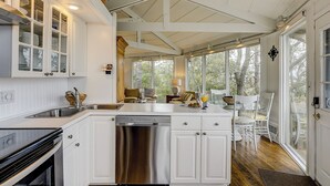 A spacious kitchen with white cabinetry, a stainless steel dishwasher, and a large island. There is a dining area with white chairs and a table, and large windows providing natural light and views of the outdoors.