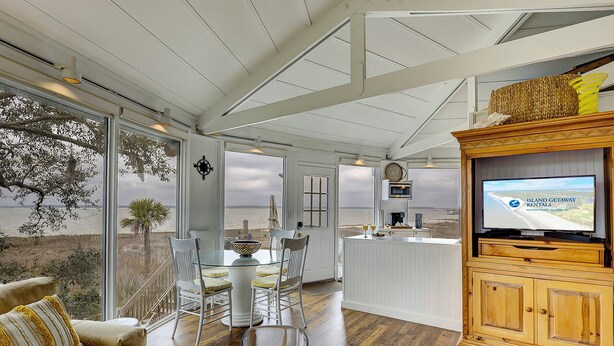 A spacious room with a large window overlooking the beach, a dining area with a glass table and chairs, a kitchen area with a TV displaying 'Island Getaway Rentals', and a wooden cabinet with a woven basket on top.