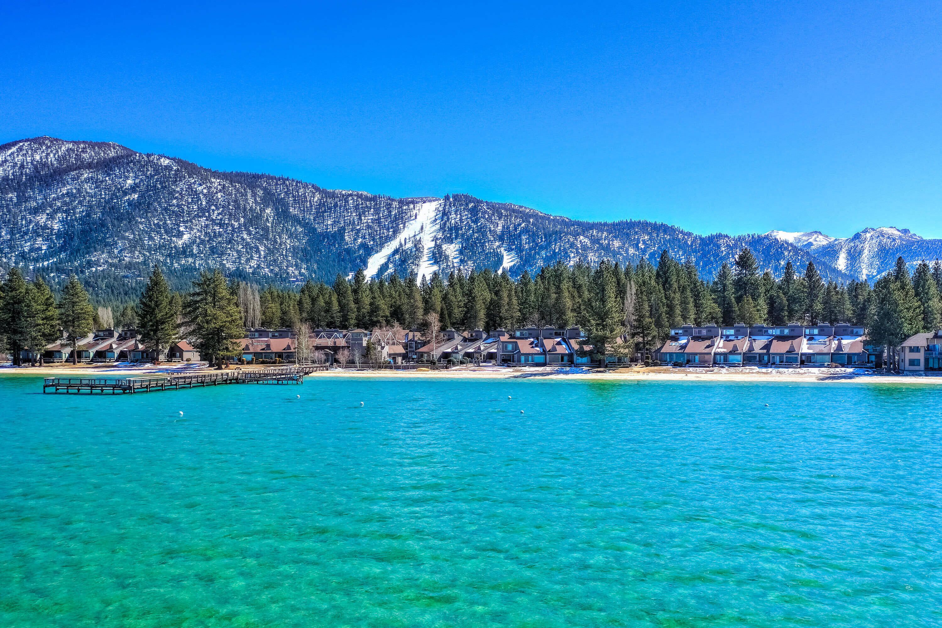 Een kustresort met een pier die zich uitstrekt over turkoenblauw water, omgeven door besneeuwde bergen en dichte bossen.