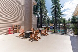 An outdoor patio with Adirondack chairs and a table, a pool in the background, and a building with horizontal siding.