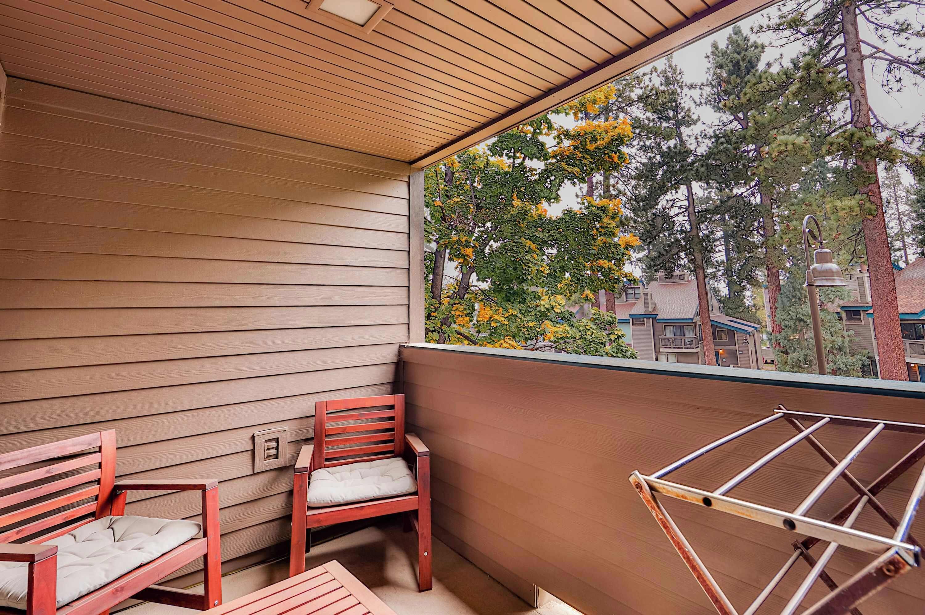 A balcony with wooden flooring, two wooden chairs, a metal drying rack, and a view of trees and buildings.