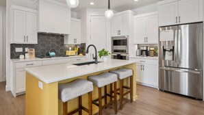 A modern kitchen with white cabinets, a large island with bar stools, a stainless steel refrigerator, and a microwave. - Beech Bungalow: Desert Color Lagoon + Beaches,  Hot Tub (St. George)