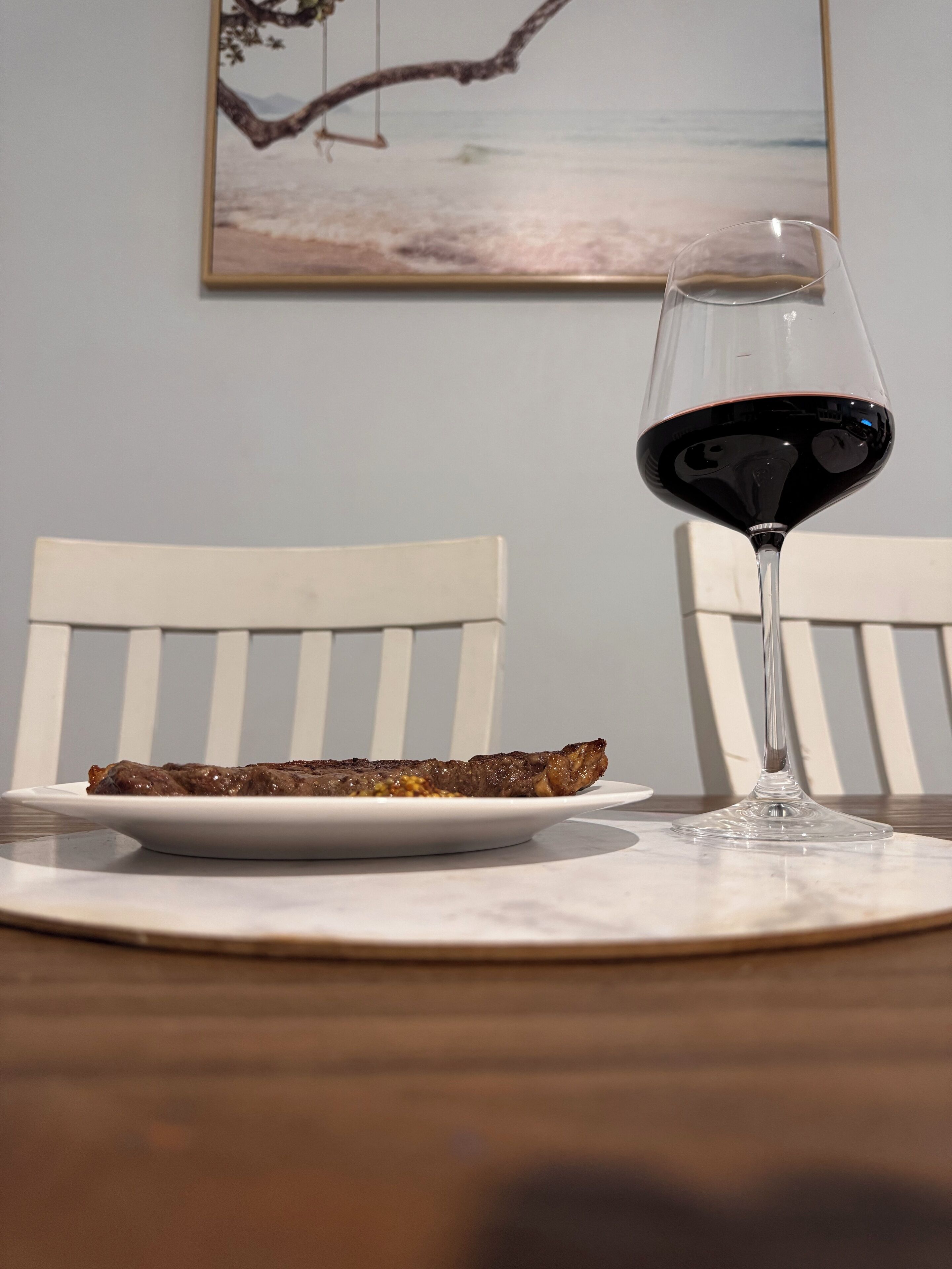 Un plato con bistec y una copa de vino tinto sobre una mesa, con una silla blanca al fondo.