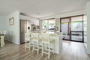 Une cuisine moderne dotée d’un îlot central, de tabourets de bar, d’un réfrigérateur et d’une porte-fenêtre coulissante donnant sur un espace extérieur.