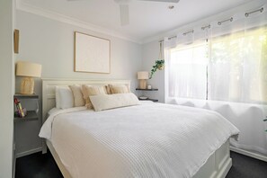 Une chambre à coucher bien rangée, avec un grand lit, des tables de chevet, une plante suspendue et une fenêtre ornée de rideaux légers.