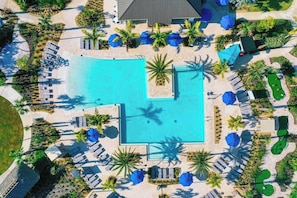 An aerial view of a resort with a central swimming pool, sun loungers, and palm trees.