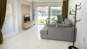 A modern living room with a grey sofa, a flat-screen TV, and a view of a balcony with outdoor furniture. - Beachfront Villa Kyma. Your own paradise (Faistos)
