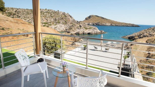 A balcony with white chairs, a small round table, and a potted plant. The balcony overlooks a rocky coastline with clear blue water and a distant hill. - Beachfront Villa Kyma. Your own paradise (Faistos)