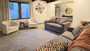 A living room with a grey sofa, two armchairs, and a patterned throw pillow. There is a window with curtains, a door, and a wall with framed pictures.