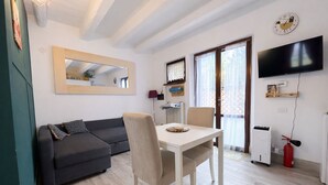 Una sala de estar moderna con un sofá gris oscuro, una mesa de comedor blanca y una silla color beige. Hay un espejo grande en la pared, un televisor de pantalla plana montado arriba y un extintor de incendios rojo en el suelo.