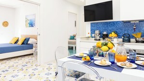 A modern hotel room with a bed, a dining area with a table set for breakfast, and a kitchen area with a TV mounted above the counter. - Marescala Vacation - Casa Cicirusso, elegant apartment in Sorrento town center (Sorrento)