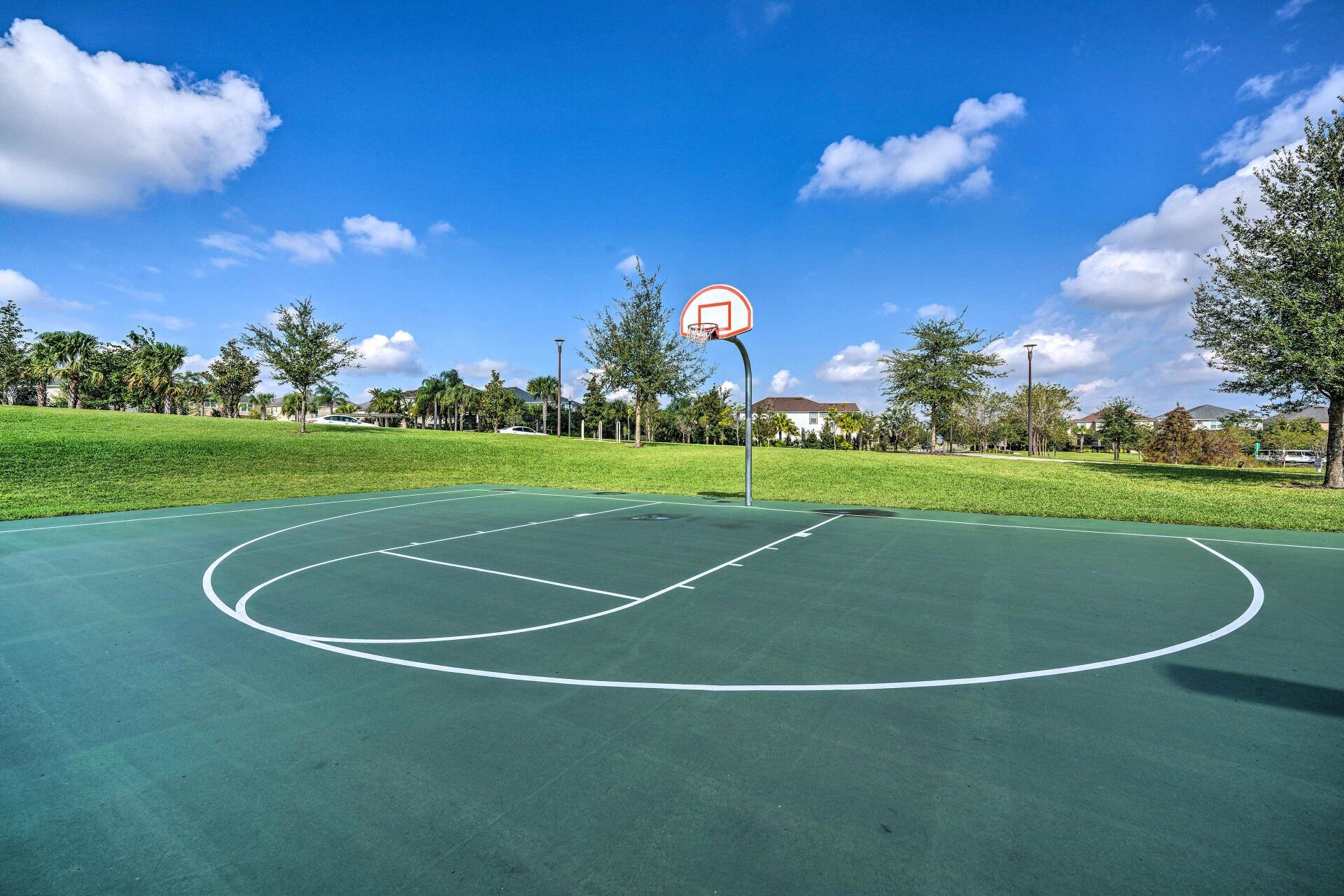 An outdoor basketball court with a hoop, surrounded by trees and a clear sky.