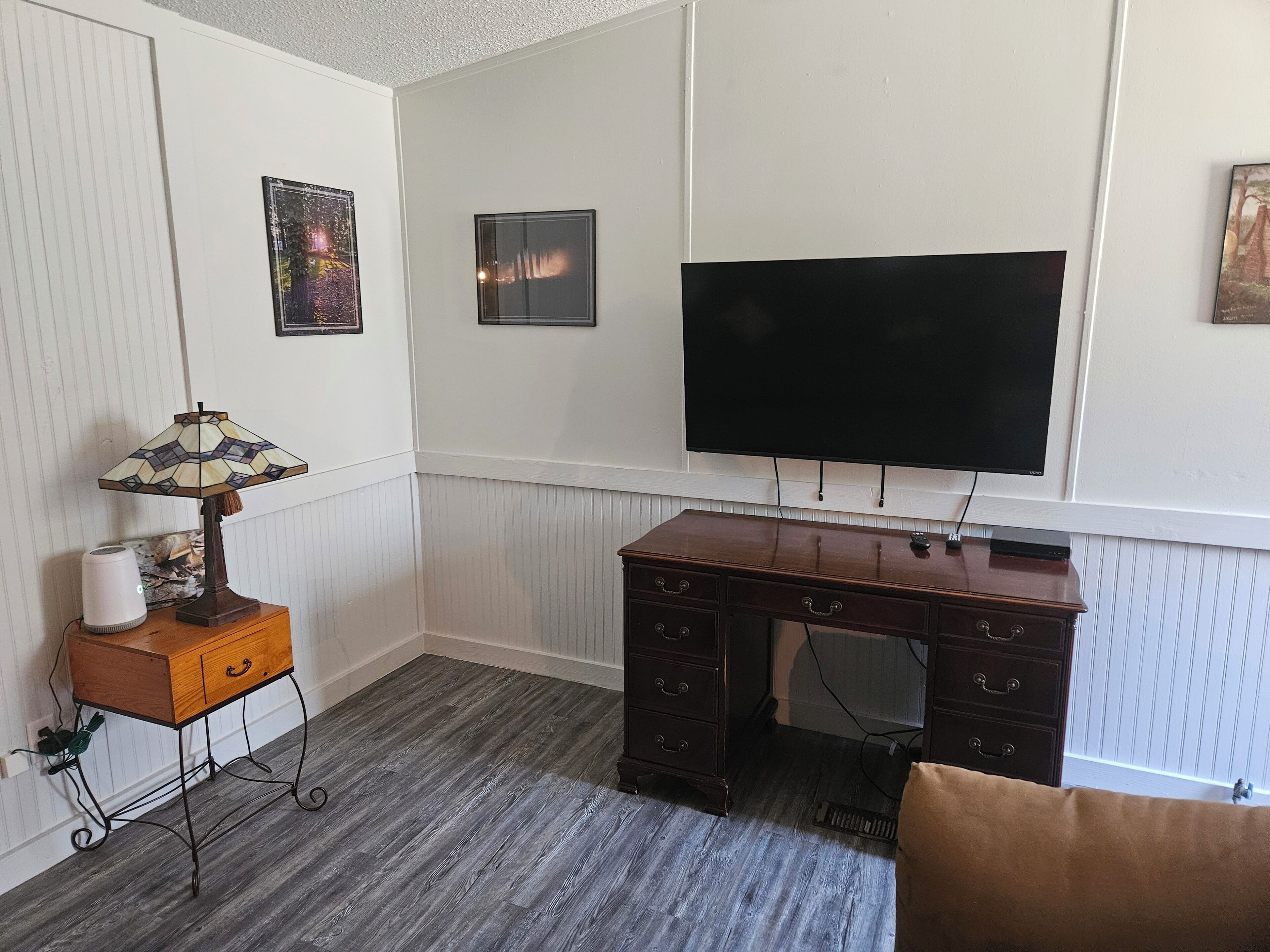 Una habitación con un escritorio de madera, un televisor de pantalla plana montado en la pared, una lámpara en una mesita auxiliar y un radiador.