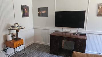 Una habitación con un escritorio de madera, un televisor de pantalla plana montado en la pared, una lámpara en una mesita auxiliar y un radiador.