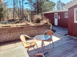 A wooden deck with two wicker chairs and a small table, surrounded by a wooden fence and a red building.