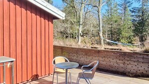 Een houten terras met een metalen tafel en twee stoelen, een rood gebouw en een houten omheining met bomen op de achtergrond.