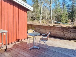 A wooden deck with a metal table and two chairs, a red building, and a wooden fence with trees in the background. - 6 Person Holiday Home in Albaek (Aalbaek)
