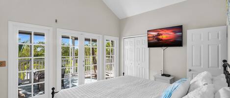 A bedroom with a bed, a TV mounted on the wall, a ceiling fan, and a view of the outdoors through large windows.