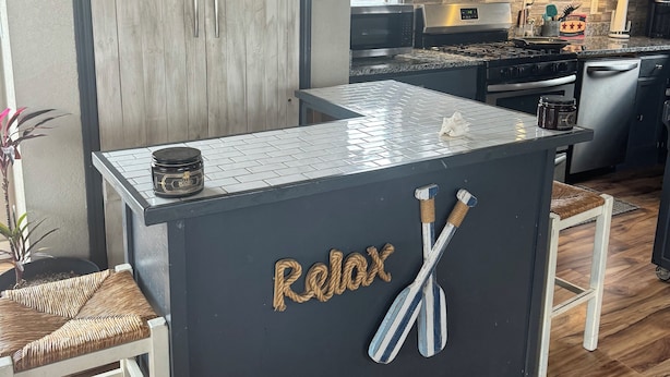 A kitchen with dark cabinets, a central island with a 'Relax' sign, and a 'Happiness' sign above the door.