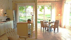 A kitchen with white cabinets, a dining area with wicker chairs, and a view of a garden through large windows. - Cottage Brantôme Swimming pool Tennis Castle Monciaux (Bourrou)