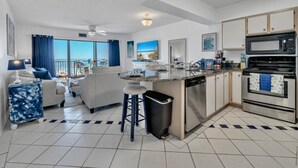Cocina moderna con una isla central, horno integrado y microondas. Cuenta con un comedor con sofá y sillas. La habitación tiene un ventanal con vistas al mar.