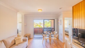 A bright living room with a sofa, a dining table with chairs, a TV, and a sliding glass door leading to a balcony with a barbecue grill. - Amanda Court 14, 1-5 Weatherly Close - 250 metres from Shoal Bay Beach (Nelson Bay)
