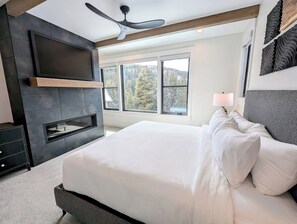 A modern bedroom with a large bed, a built-in TV, a fireplace, and large windows offering a view of the outdoors. - Luxury | Hot Tub | Walk to River Run Gondola (Keystone)