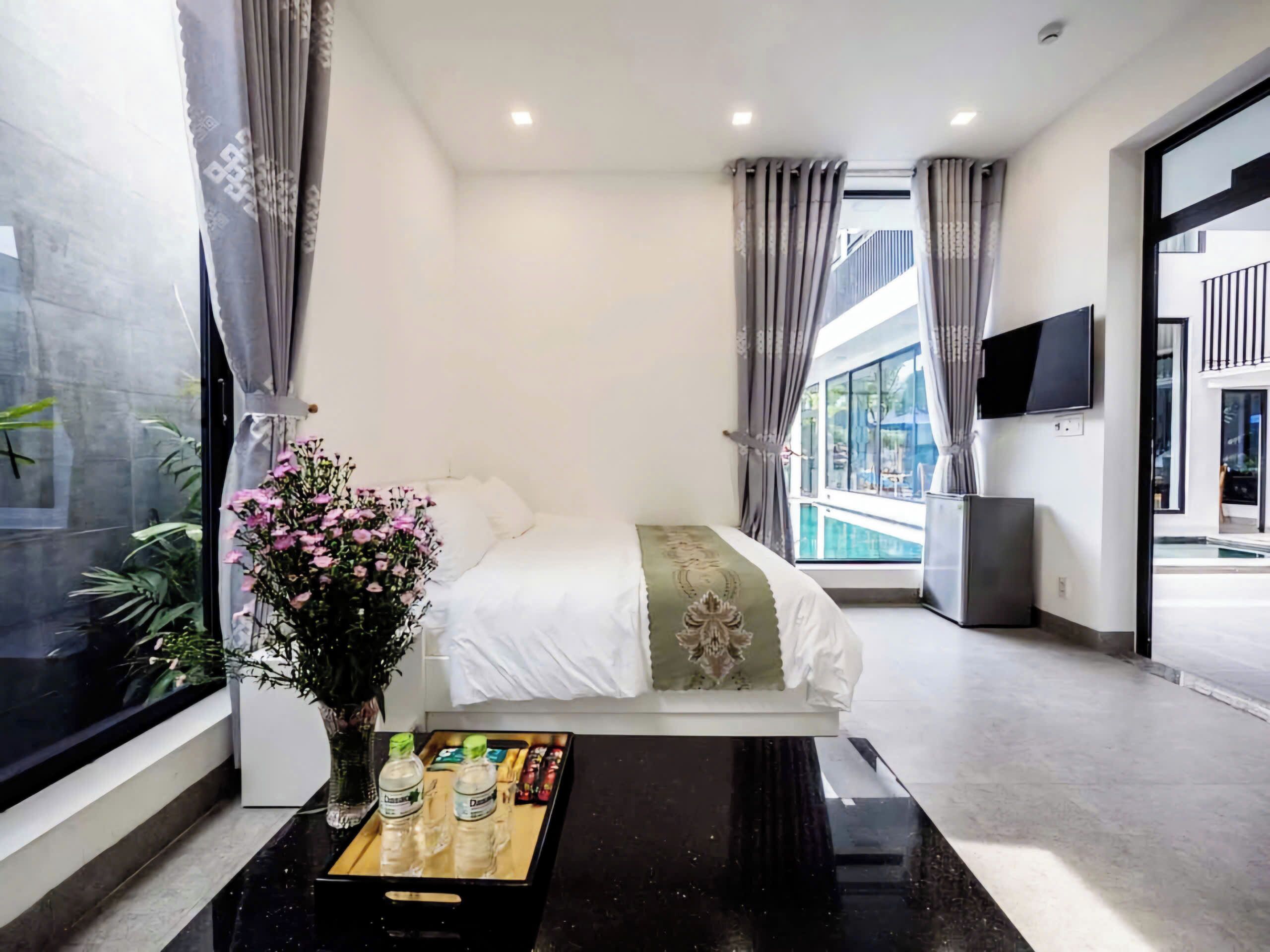Een moderne hotelkamer met een groot bed, een nachtkastje met een bloevas, een flatscreen televisie aan de muur en uitzicht op een zwembad door het raam.