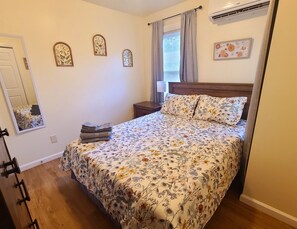 A bedroom with a floral bedspread, a wooden headboard, a bedside table with a lamp, and a window with curtains.