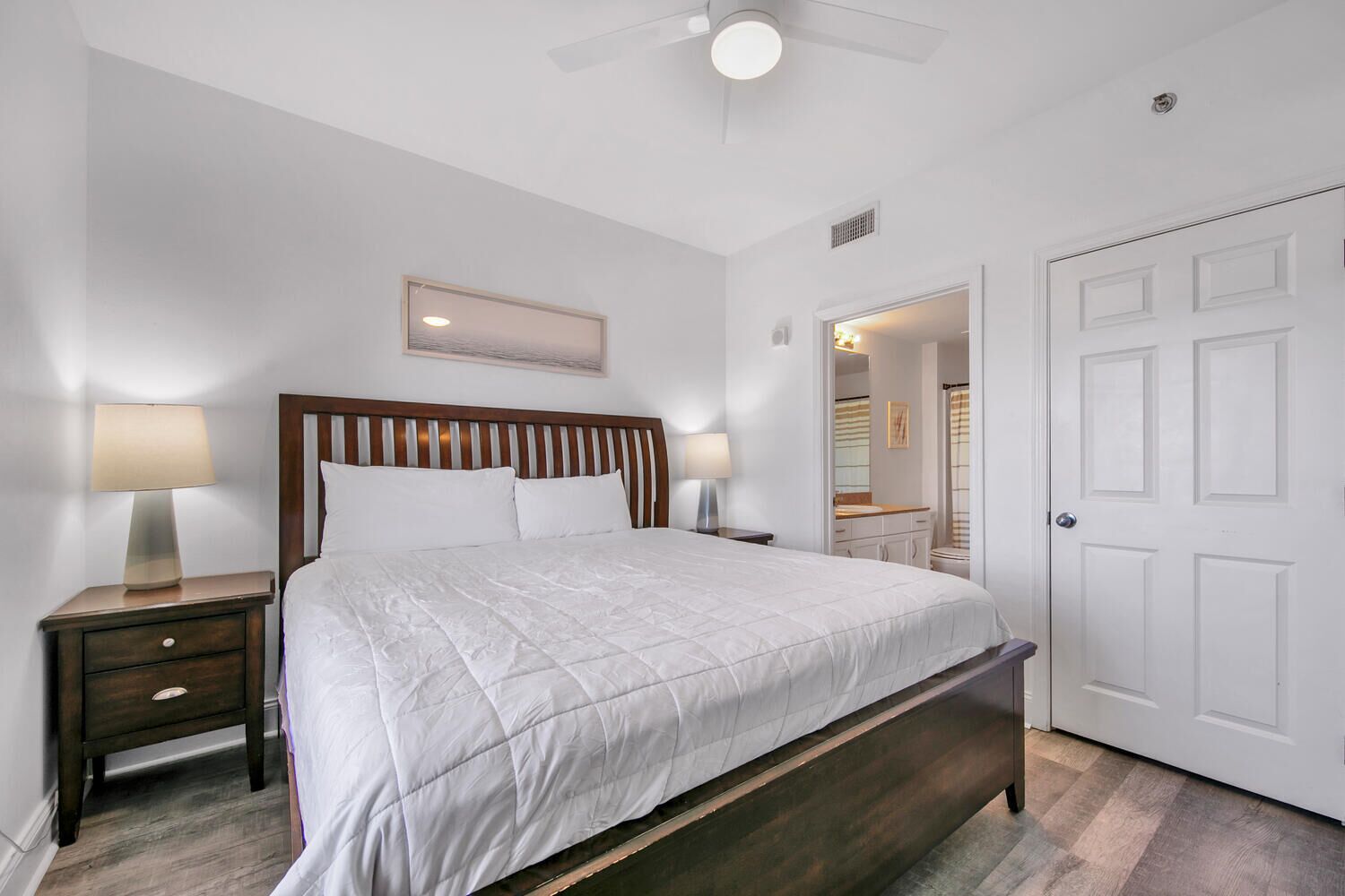 A bedroom with a large bed, two bedside tables with lamps, a ceiling fan, and a white door.