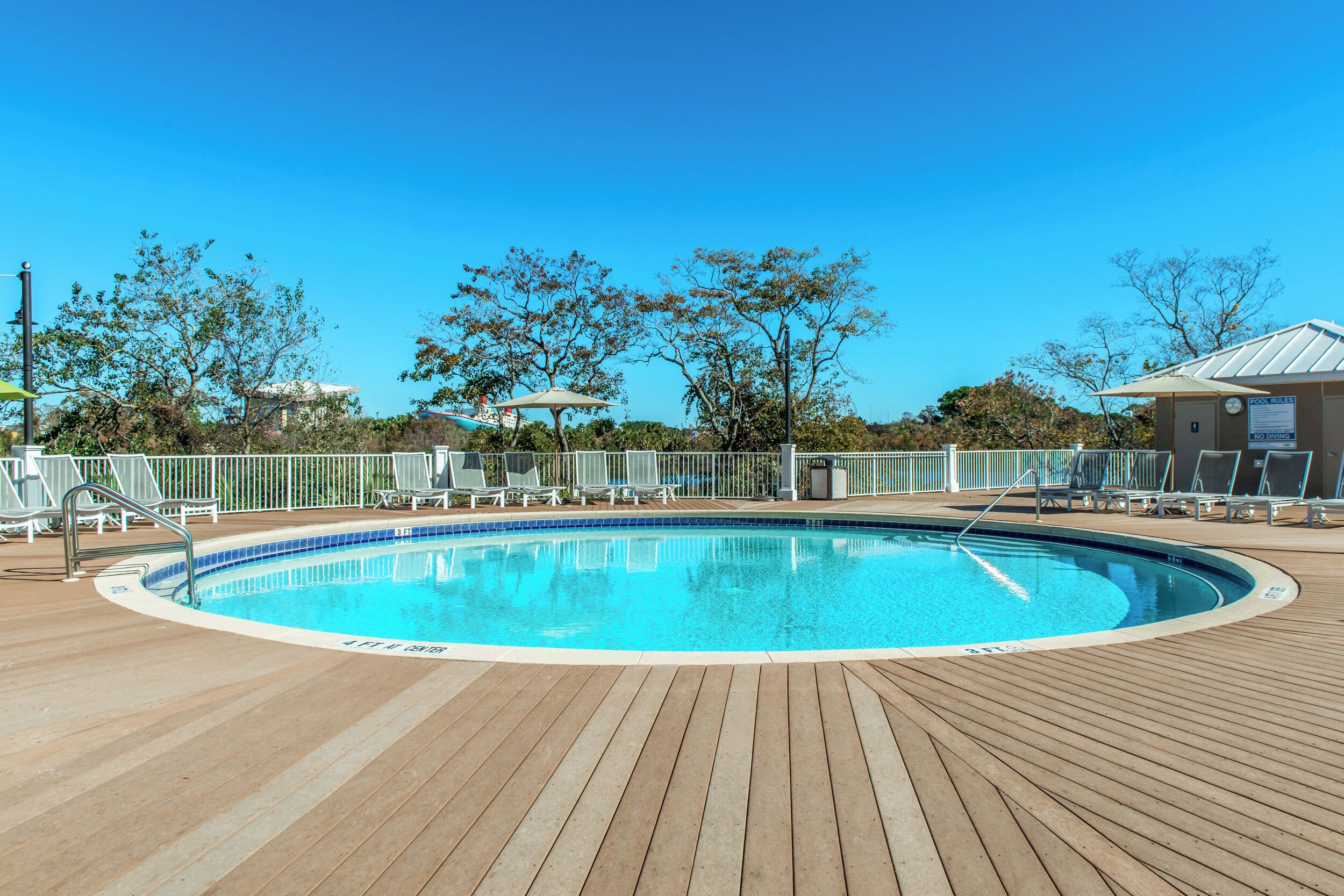 A wooden deck with a swimming pool, lounge chairs, and a fenced area with trees.