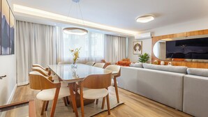 Ein modernes Wohnzimmer mit Essbereich, ausgestattet mit einem Holzesstisch, weißen Stühlen, einem grauen Sofa und einem an der Wand befestigten Flachbildfernseher.