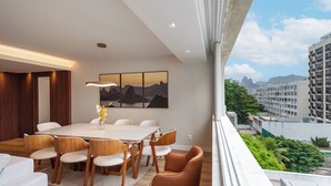 Ein modernes Hotelzimmer mit großem Esstisch, bequemen Sitzgelegenheiten, Blick auf die Stadt und einem Balkon mit Grünpflanzen.