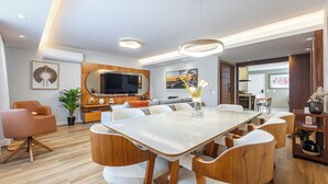 Ein modernes Wohnzimmer mit großem Esstisch, bequemen Sitzgelegenheiten, einem Flachbildfernseher und einer im Hintergrund sichtbaren Küchenzeile.