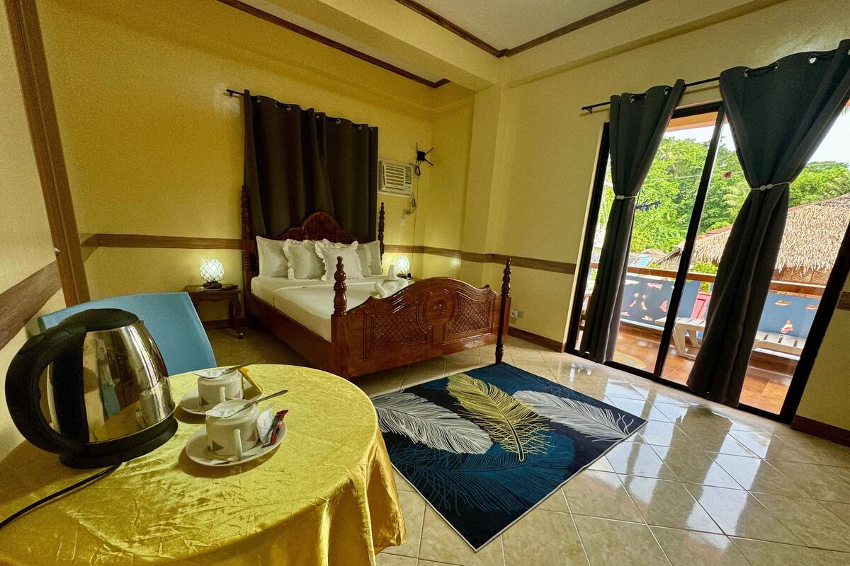 Habitación de hotel con cama, una mesa con hervidor y tazas, una alfombra azul, y vistas al exterior a través de un ventanal.