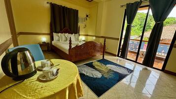A hotel room with a bed, a table with a kettle and cups, a blue rug, and a view of the outdoors through a glass door.