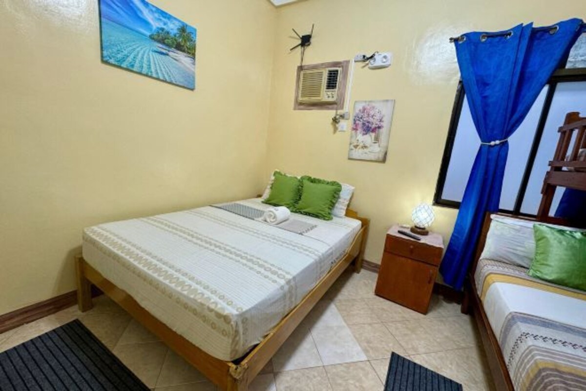 A bedroom with a bed, a nightstand, a lamp, a window with blue curtains, and a wall-mounted air conditioner.