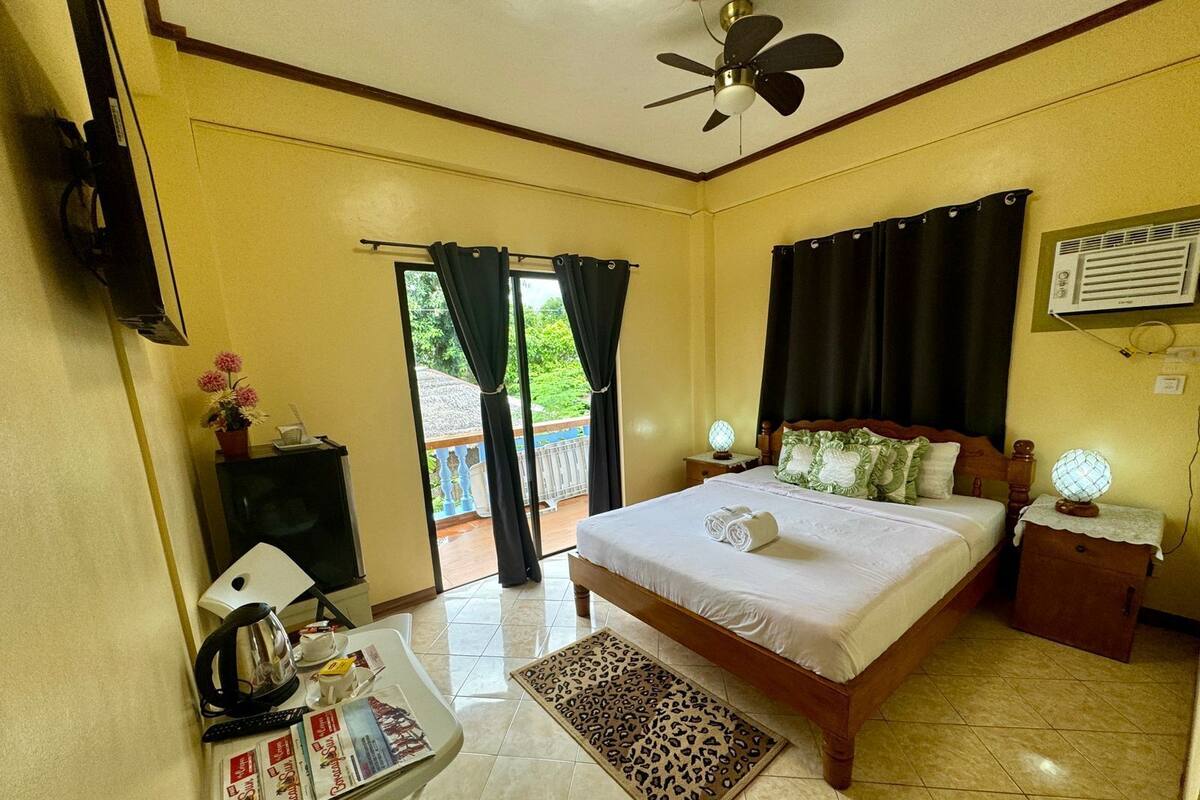 Habitación de hotel con cama, escritorio con silla, televisor, ventilador de techo y ventana con cortinas.
