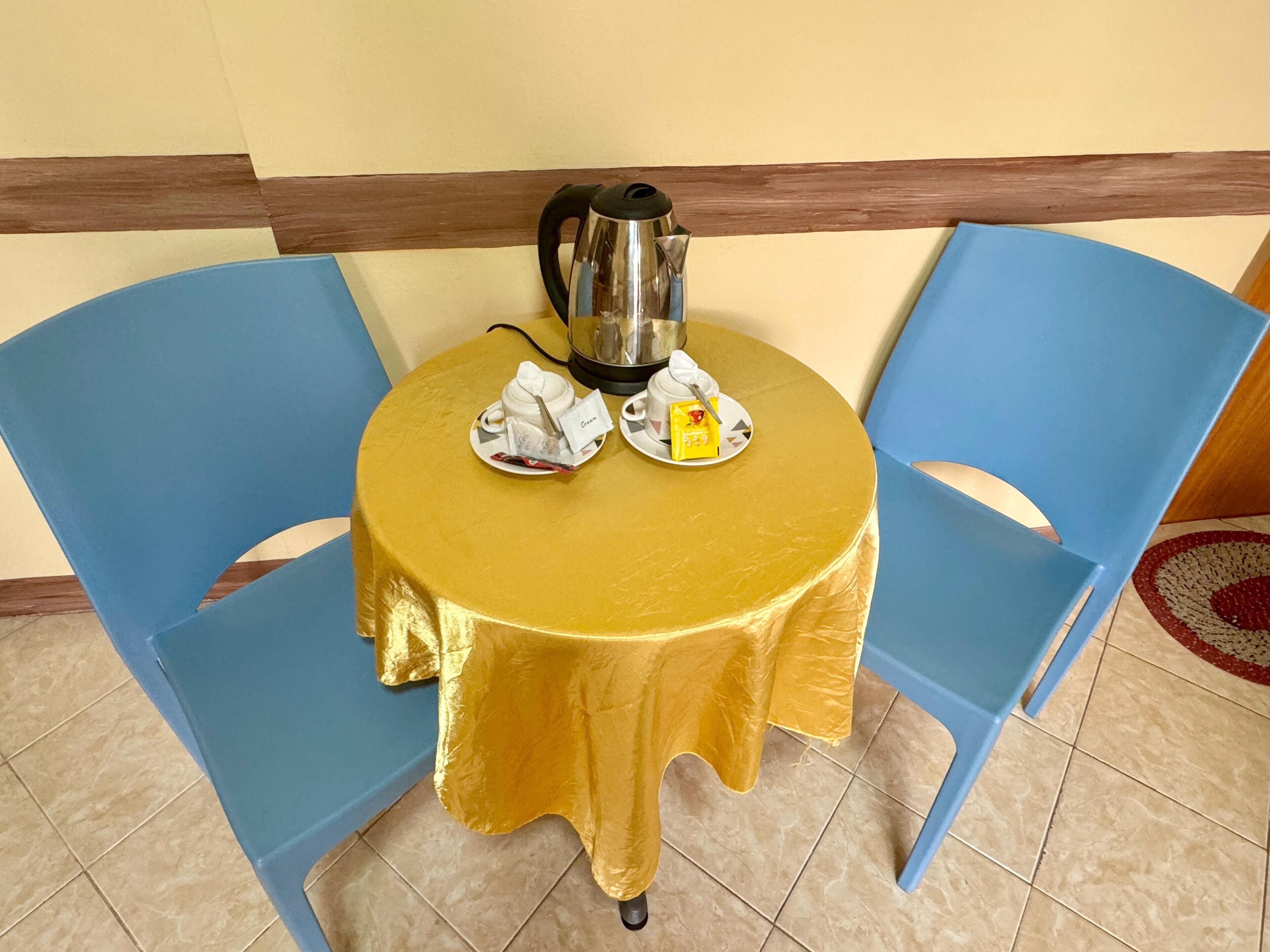 A round table with a gold tablecloth, two blue chairs, a kettle, and a small plate with a teabag and a packet.