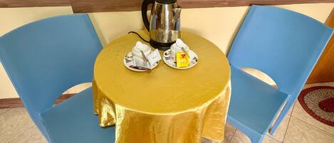 A round table with a gold tablecloth, two blue chairs, a kettle, and a small plate with a teabag and a packet.