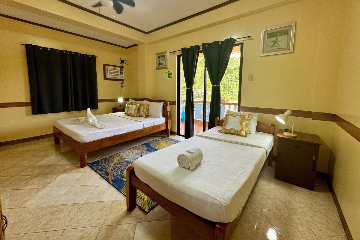 Habitación de hotel con dos camas individuales, un ventilador de techo, una mesita de noche con lámpara y vistas al exterior a través de una ventana con cortinas.
