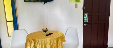 A room with a television mounted on the wall, a table with a yellow tablecloth, two white chairs, and a brown door.