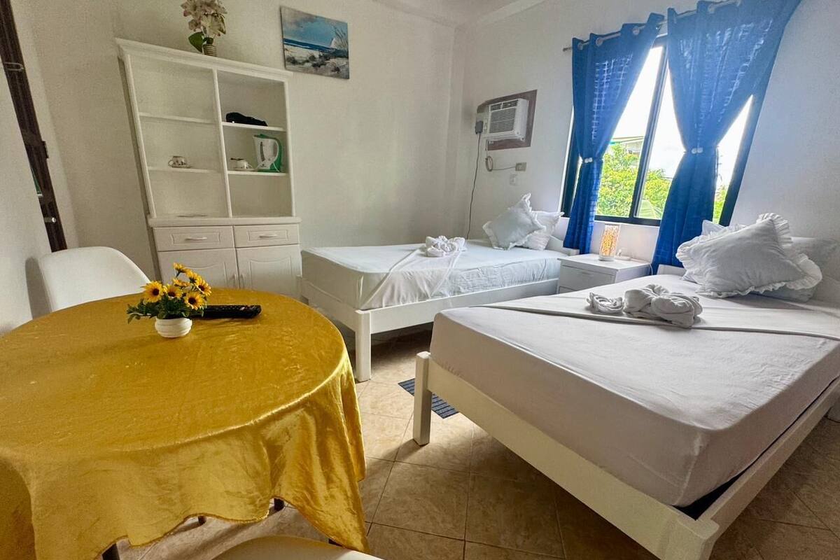 Habitación de hotel con dos camas, una mesa de comedor con mantel amarillo, un armario blanco y una ventana con cortinas azules.
