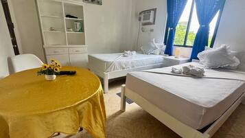 A hotel room with two beds, a dining table with a yellow tablecloth, a white wardrobe, and a window with blue curtains.