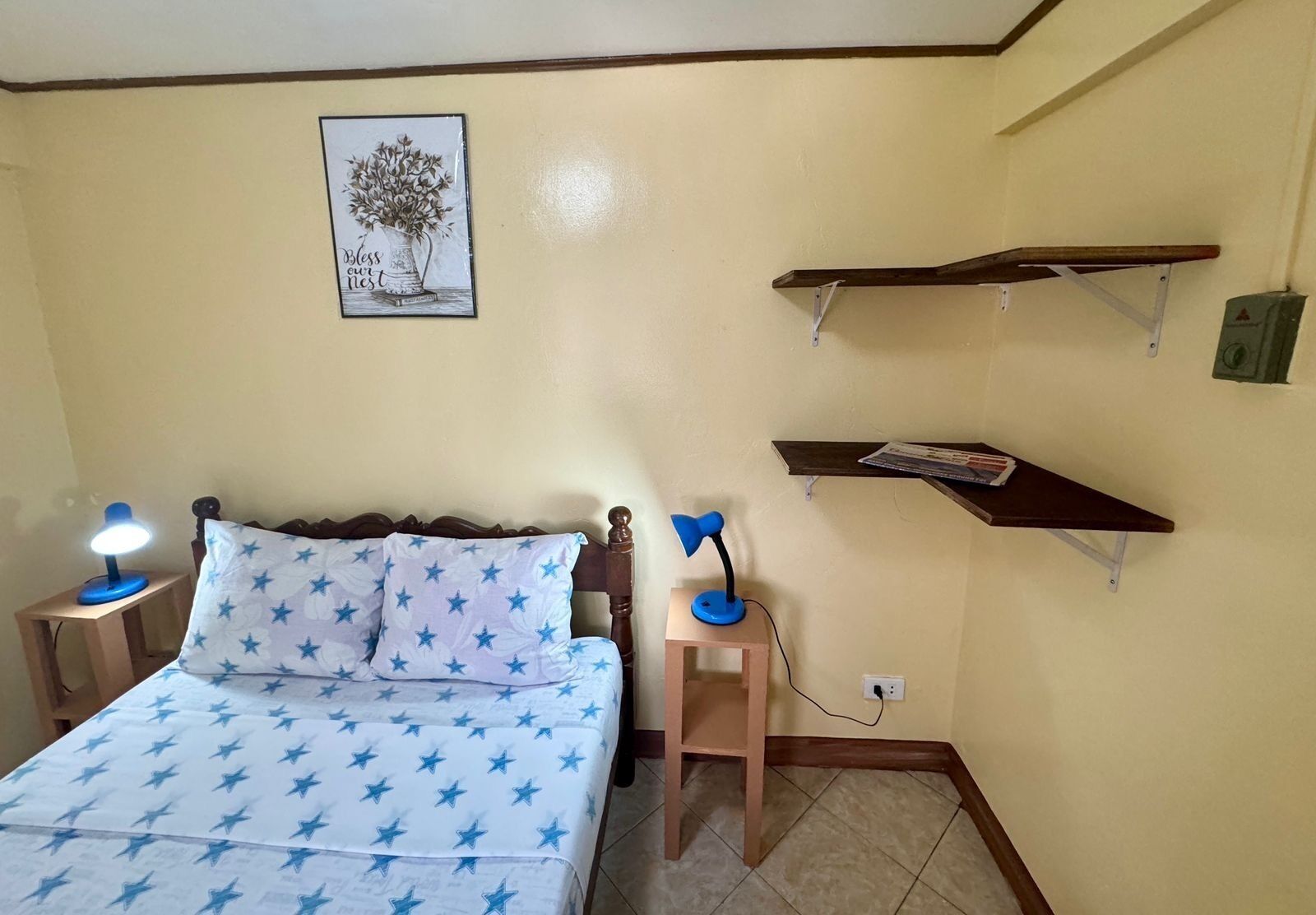 A bedroom with a bed, two bedside lamps, a nightstand, a wall-mounted shelf, and a framed picture.