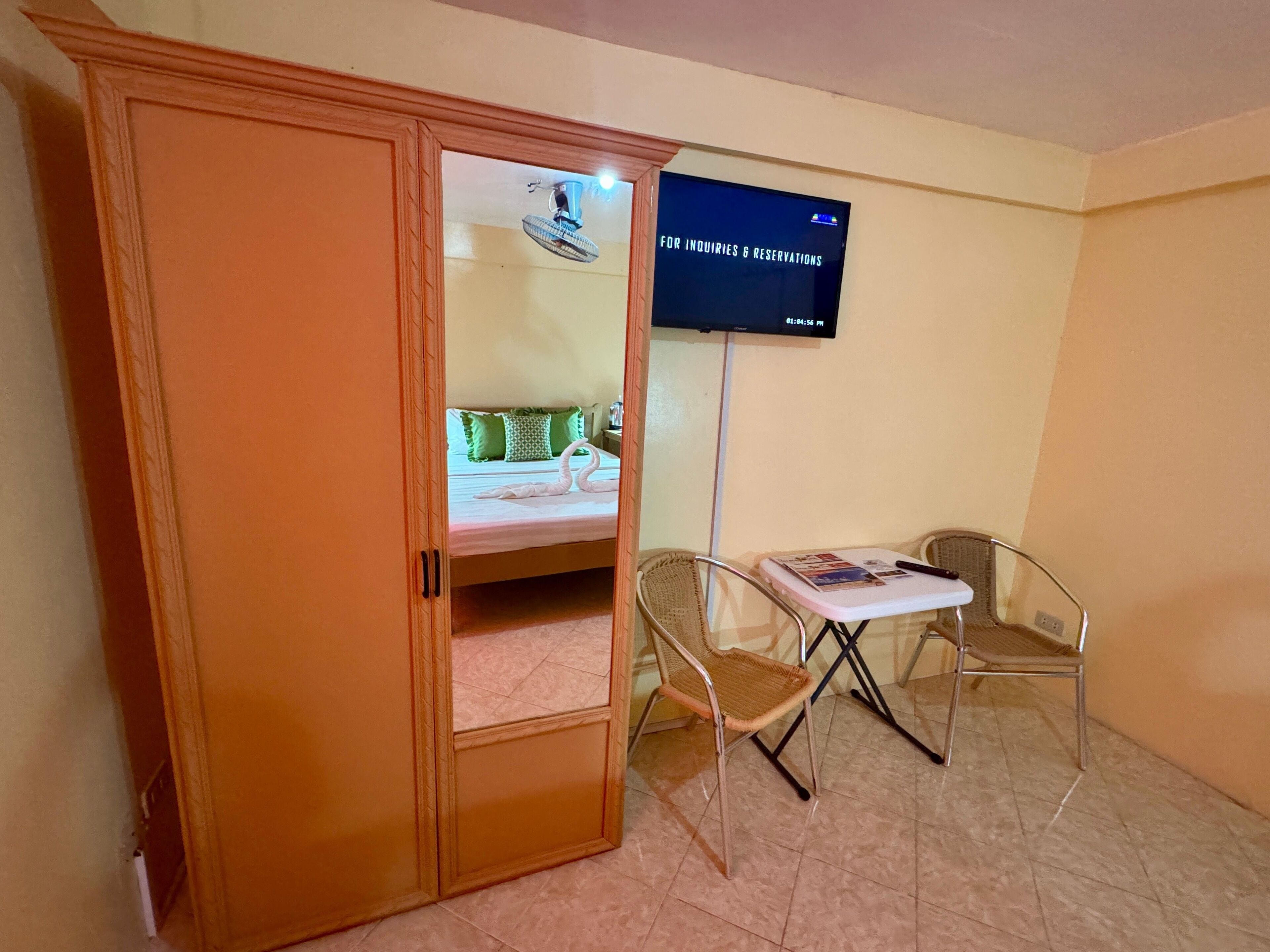 A hotel room with a wardrobe, a bed with green pillows, a wall-mounted TV, and two chairs.