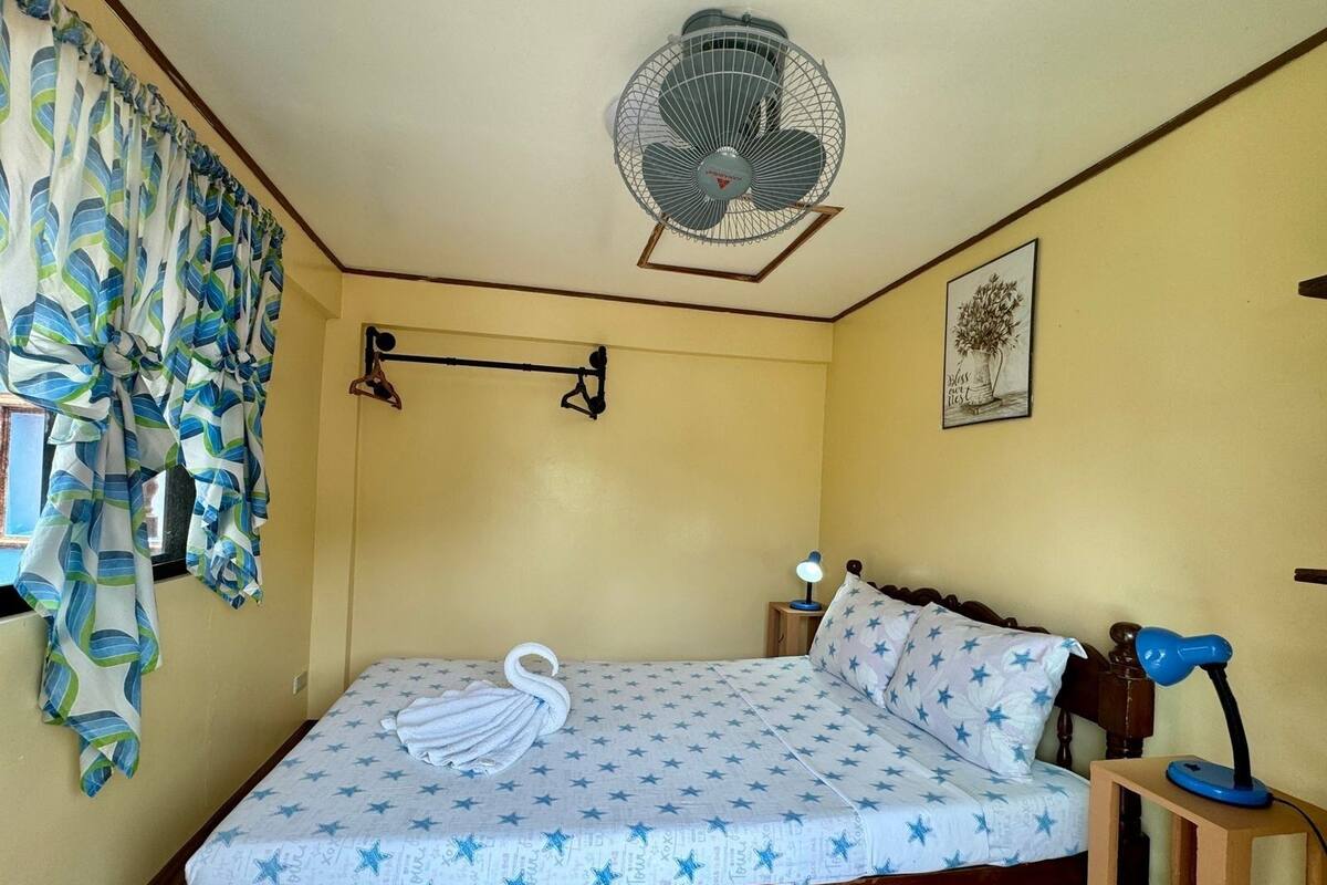 Un dormitorio con cama, ventilador de techo, mesita de noche con lámpara y ventana con cortinas.