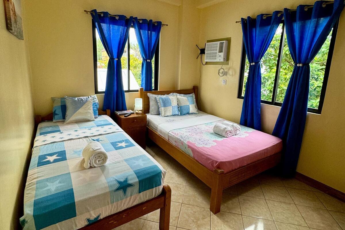 Una habitación con dos camas, un ventilador de techo y ventanas con cortinas azules.