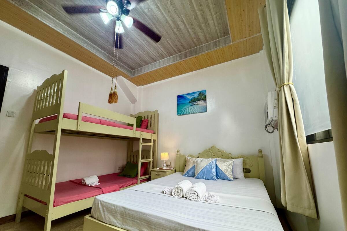 Una habitación con una cama litera, una cama individual, un ventilador de techo, un cuadro y una mesita de noche con lámpara.