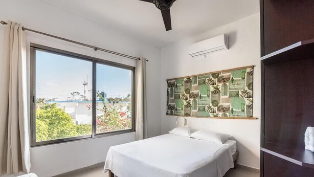 A bedroom with a large window, a bed, a ceiling fan, and a wall-mounted air conditioner. - Beautiful complete 2 bedroom apartment, Tulum Centro! Aluxe (Tulum)